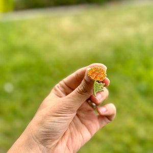 May include: A gold-tone enamel pin featuring a marigold flower with orange petals and green leaves.