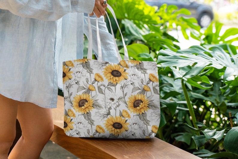 K&ouml;nnte beinhalten: Eine wei&szlig;e Einkaufstasche mit einem Muster aus Sonnenblumen und wei&szlig;en Blumen. Die Tasche hat wei&szlig;e Henkel und ist aus Canvas.
