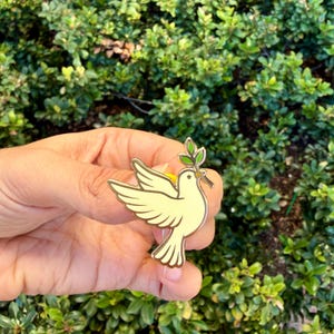 May include: A gold-colored dove pin with a light yellow body and a green olive branch in its beak. The pin has a detailed design with a gold outline. The background is a lush green bush.