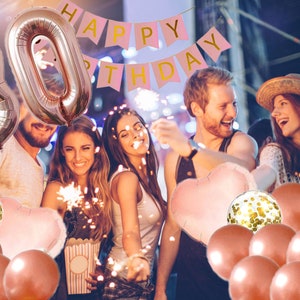 May include: A group of friends celebrating a 30th birthday with rose gold balloons, confetti balloons, and a pink banner that says "Happy Birthday".