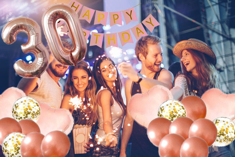 May include: A group of friends celebrating a 30th birthday with rose gold balloons, confetti balloons, and a pink banner that says "Happy Birthday".