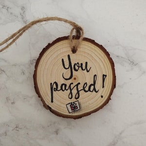 May include: A round wooden ornament with the text "You passed!" written in black ink. The ornament is attached to a piece of twine.