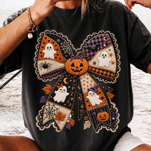 Può includere: Una t-shirt nera con una grande grafica centrale a forma di fiocco realizzata con vari motivi in tessuto a tema Halloween. Il design include fantasmi bianchi, zucche arancioni, ragnatele, pipistrelli e foglie autunnali con un bordo smerlato.