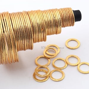 Puede incluir: Una pila de anillos de metal de color dorado, con algunos anillos dispersos sobre una superficie blanca. Los anillos son lisos y tienen un acabado ligeramente texturizado.