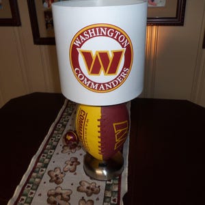 Puede incluir: Una pantalla de lámpara blanca con un logotipo de los Washington Commanders en rojo y dorado. La base de la lámpara es una pelota de fútbol con el logotipo y los colores del equipo.