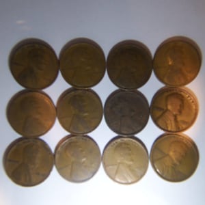 May include: Twelve copper-colored pennies arranged in three rows of four. The coins show the profile of a person on one side. The pennies are in varying conditions, with some appearing more worn than others.