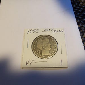 May include: An 1895 silver half dollar coin, displayed in a protective holder. The coin features a profile of a woman and the text "IN GOD WE TRUST". The holder is labeled "1895 HALF DOLLAR" and "VF".