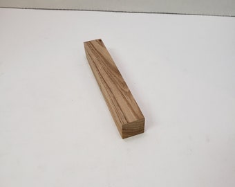 May include: A rectangular block of light brown wood with a visible grain pattern. The wood block is a solid, square shape, likely for crafting or woodworking projects. The wood appears to be unfinished.