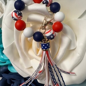 May include: A patriotic keychain wristlet featuring a ring of red, white, and blue beads. The keychain has a gold clasp and a tassel with red, white, and blue striped and star-printed accents. The wristlet is set against a backdrop of white and blue floral decorations.