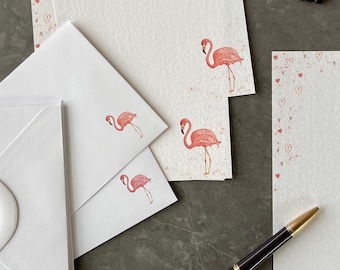 May include: A set of pink flamingo stationery with envelopes. The paper has a light pink background with a scattering of small pink hearts. The envelopes are white with a pink flamingo illustration.