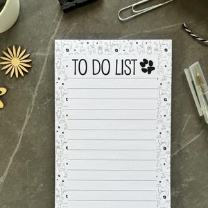 May include: A white to-do list notepad with a black paw print and the text "TO DO LIST" at the top. The notepad has a black and white pattern of paw prints and bones around the edges. A clear pen is lying on a gray surface next to the notepad.