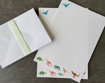 May include: A set of white stationery with a blue bird design on the top and a border of colorful dinosaurs on the bottom. The stationery is folded in half and has a light green ribbon.