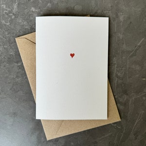 May include: A blank white card with a small red heart printed in the center. The card is sitting on a brown envelope.