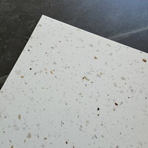 Puede incluir: Primer plano de una superficie blanca moteada con peque&ntilde;as motas marrones y doradas. La superficie es lisa y tiene una textura sutil.