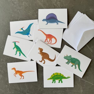 May include: Set of 8 blank greeting cards featuring different dinosaur illustrations. The cards are white with colourful dinosaur illustrations, including a stegosaurus, a pterodactyl, a triceratops, and a tyrannosaurus rex.