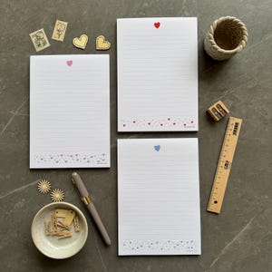 May include: Three lined white notepads with a heart at the top and a heart border at the bottom. A pen, pencil sharpener, ruler, and small bowl with mini clothespins are also present. Decorative stamps and wooden hearts are scattered around.
