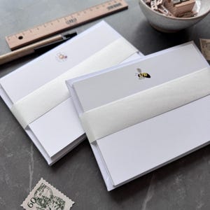 May include: Two stacks of white stationery, each secured with a white band. One features a bee illustration, the other a daisy. A wooden ruler and pencil are in the background, with a bowl of stamps and a postage stamp.