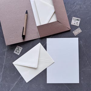 May include: A stationery set featuring a brown folder, cream-colored envelopes, and a lined white paper. A pen and two vintage postage stamps are also included. The set is arranged on a dark gray surface.