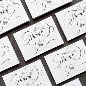 May include: A collection of rectangular white greeting cards featuring the words Thank You printed in a black, elegant, flowing cursive script. The cards are arranged in a scattered pattern, showcasing the decorative calligraphy style.