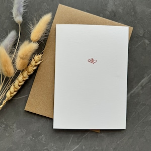 May include: A white greeting card featuring two small, interconnected red hearts printed in the center. The card is placed on top of a brown kraft paper envelope, accompanied by a small arrangement of dried beige and light purple grasses.