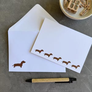 May include: A white stationery set consisting of a blank note card and a matching envelope. The note card features a row of five brown dachshund illustrations with colourful collars, while the envelope displays a single brown dachshund.
