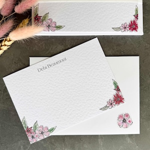 May include: A white card with the name "Debi Beaumont" printed in black ink. The card features a floral design in pink and red on the bottom corner. The card is sitting on top of a white envelope with a similar floral design on the bottom corner.