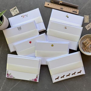 May include: A collection of white stationery note cards bundled with cream paper bands, featuring various small illustrations including cocktails, a bee, a daisy, a bow, a red heart, a yellow star, pink flowers, and a row of brown dachshund dogs.