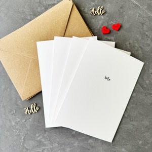 May include: Four blank white cards with the word "hello" written on one of the cards. The cards are stacked on top of each other and are sitting on a brown paper envelope. There are two red heart-shaped buttons on the table.