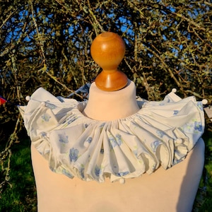 May include: A white ruffled collar with a blue floral pattern. The collar is on a dress form with a wooden head.