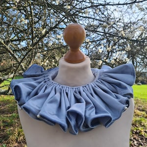 Könnte beinhalten: Ein hellblauer Rüschenkragen aus Samt auf einer Schaufensterpuppe. Der Kragen hat ein gerafftes Design und ist mit kleinen, weißen Perlen verziert. Der Hintergrund zeigt eine verschwommene Außenszene mit Bäumen und grünem Gras.