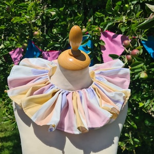 May include: A ruffled neck collar featuring vertical stripes in pastel shades of pink, yellow, and lavender. The fabric is gathered into dense, voluminous folds around the neckline, accented with small gold-toned beads at the outer edges.