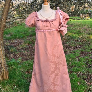 May include: A dusty rose-colored empire waist gown with a floral damask pattern. The dress features short, puffed sleeves, a square neckline with ruffle trim, and a matching ribbon sash. A pearl necklace and a small matching purse complete the ensemble.