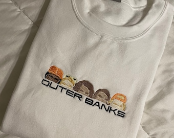 Könnte beinhalten: Weißer Rundhals-Pullover mit einem gestickten Design aus fünf Comic-Gesichtern und dem Text "OUTER BANKS".