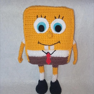 May include: A crocheted SpongeBob SquarePants toy with a yellow body, white shirt, brown trousers, and a red tie. The toy has blue eyes, a white smile, and a brown nose.