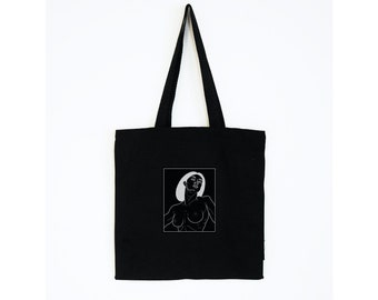 May include: Black tote bag featuring a white line drawing of a person with a hat. The tote bag has a rectangular design with a long handle. The image is centered on a white background.