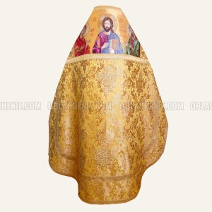 May include: Gold-colored liturgical vestment with a detailed floral pattern. The upper portion features an embroidered religious icon with figures. The vestment has a rounded shape and is trimmed with a gold border. The fabric has a shiny appearance.