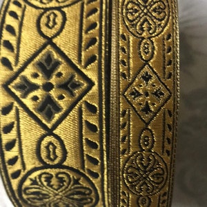 May include: Two gold and black patterned trim ribbons. The ribbon on the bottom has a more intricate design with a diamond shape in the center. The ribbon on top has a more simple design with a circular shape in the center.