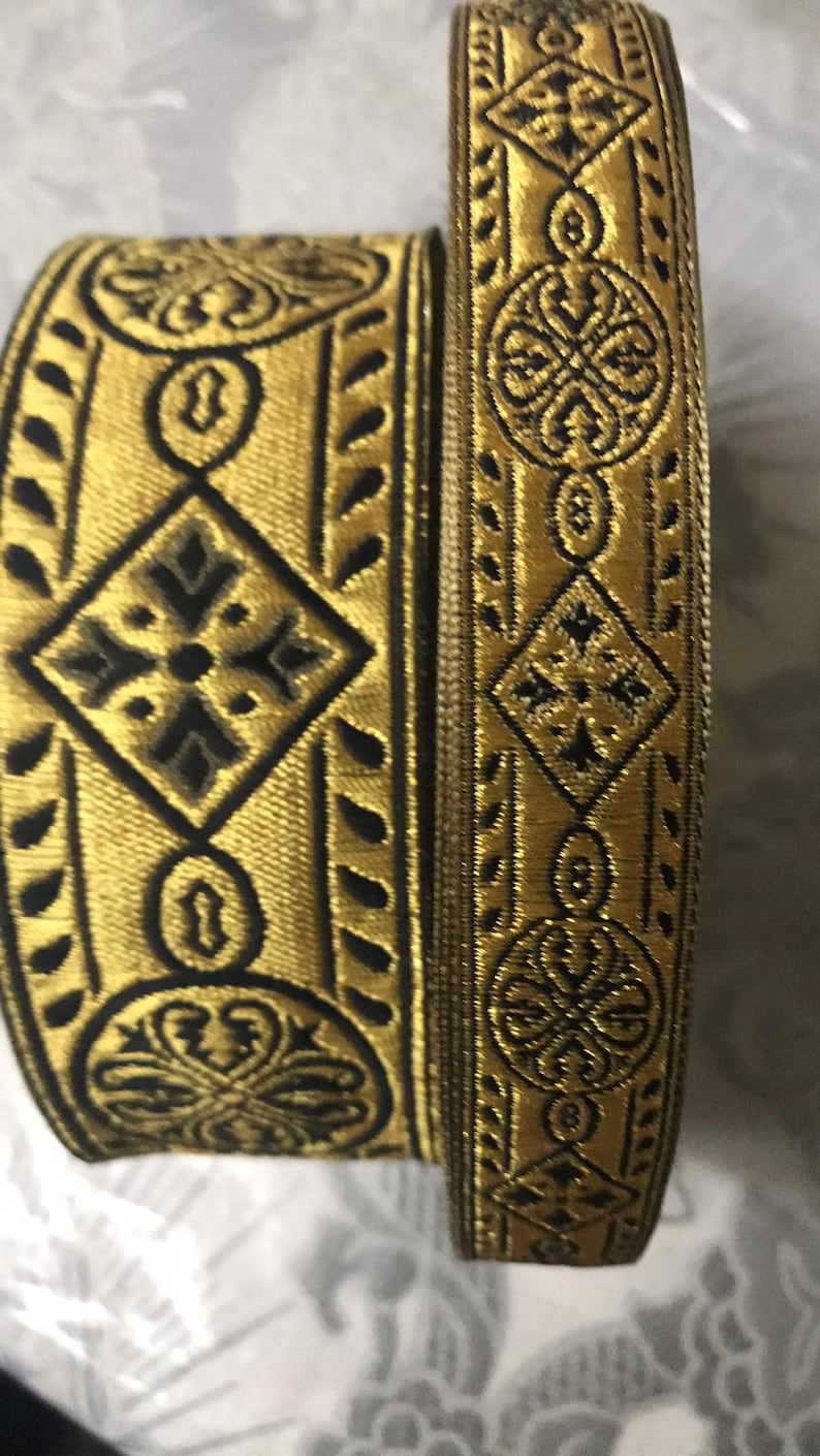 May include: Two gold and black patterned trim ribbons. The ribbon on the bottom has a more intricate design with a diamond shape in the center. The ribbon on top has a more simple design with a circular shape in the center.