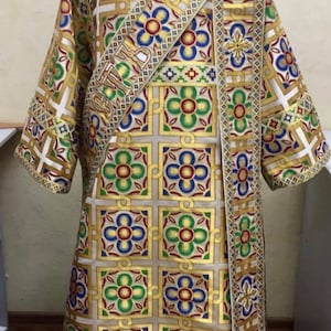 May include: A white liturgical vestment with a gold, red, green, and blue geometric pattern. The vestment has a gold sash with a cross design and a gold fringe.