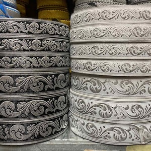 Puede incluir: Dos rollos de ribete decorativo con un patrón floral plateado y negro. El ribete está hecho de una tela tejida y tiene un diseño en relieve.