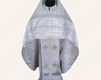 May include: A white Orthodox vestment featuring a structured phelonion with a geometric pattern and a matching sticharion. The garment is adorned with silver-toned trim, embroidered cross motifs, and decorative fringe along the bottom hem.