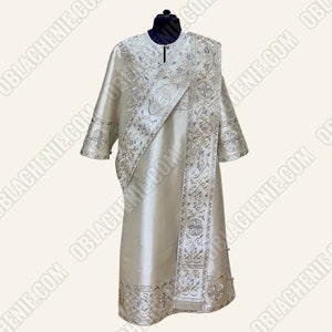 May include: A white liturgical vestment with silver embroidery. The vestment has a long, flowing robe with a wide, decorative collar. The sleeves are long and have a decorative cuff. The vestment is decorated with a silver embroidered cross and other designs.