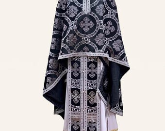 May include: A black and silver liturgical vestment set featuring a long robe and a cape. The fabric is adorned with intricate silver cross patterns and circular motifs. The garment includes a vertical central panel with silver trim and tassels.