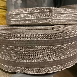 Puede incluir: Dos rollos de ribete metálico plateado con un patrón de rayas. El ribete está hecho de un material suave y flexible y tiene un brillo sutil.