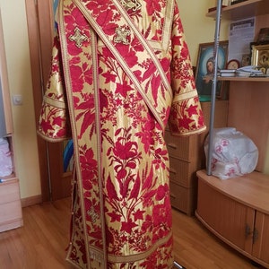Pode incluir: Uma veste litúrgica vermelha e dourada com bordados florais e uma faixa dourada com cruzes.