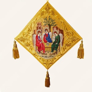 Op de afbeelding: Een ruitvormige wandhanger van goudkleurige stof met een geborduurde religieuze scène van drie figuren met halo's die aan een tafel zitten. De rand is versierd met gouden draadkrullen en drie gouden kwastjes hangen aan de hoeken.