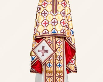 May include: A liturgical vestment set featuring a white and gold patterned fabric with repeating red and blue cross motifs. The garment includes a long robe with vertical panels, a wide cape, and a square-shaped accessory with a central cross.