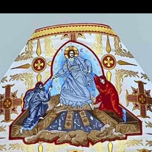 May include: A white fabric with gold embroidery depicting a religious scene. A figure in blue robes with a halo is surrounded by two figures in red and blue robes. The scene is set against a background of gold crosses and floral motifs.
