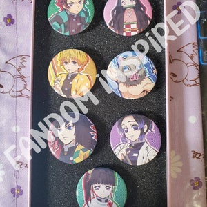 May include: A set of eight round button pins featuring characters from the anime series Demon Slayer. The pins are arranged in a grid pattern on a black background. The characters include Tanjiro Kamado, Nezuko Kamado, Zenitsu Agatsuma, Inosuke Hashibira, Giyu Tomioka, Shinobu Kocho, and Kanao Tsuyuri.
