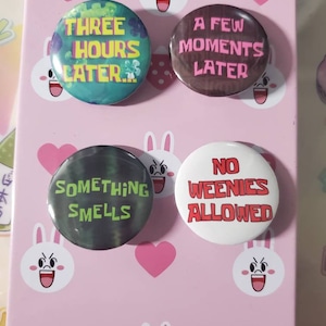 May include: Four round buttons with different sayings on them. The buttons are on a pink background with white bunny faces. The sayings on the buttons are "Three Hours Later", "A Few Moments Later", "Something Smells", and "No Weenies Allowed".